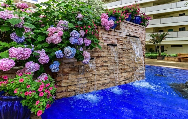 A stone wall with cascading water feature, surrounded by vibrant hydrangea flowers and petunias.
