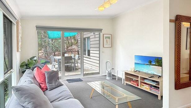 A living room with a sofa, a glass coffee table, a TV on a wooden stand, and a sliding glass door leading to an outdoor patio. - Christensen - Pet Friendly home at the base of Mt Buller (Sawmill Settlement)