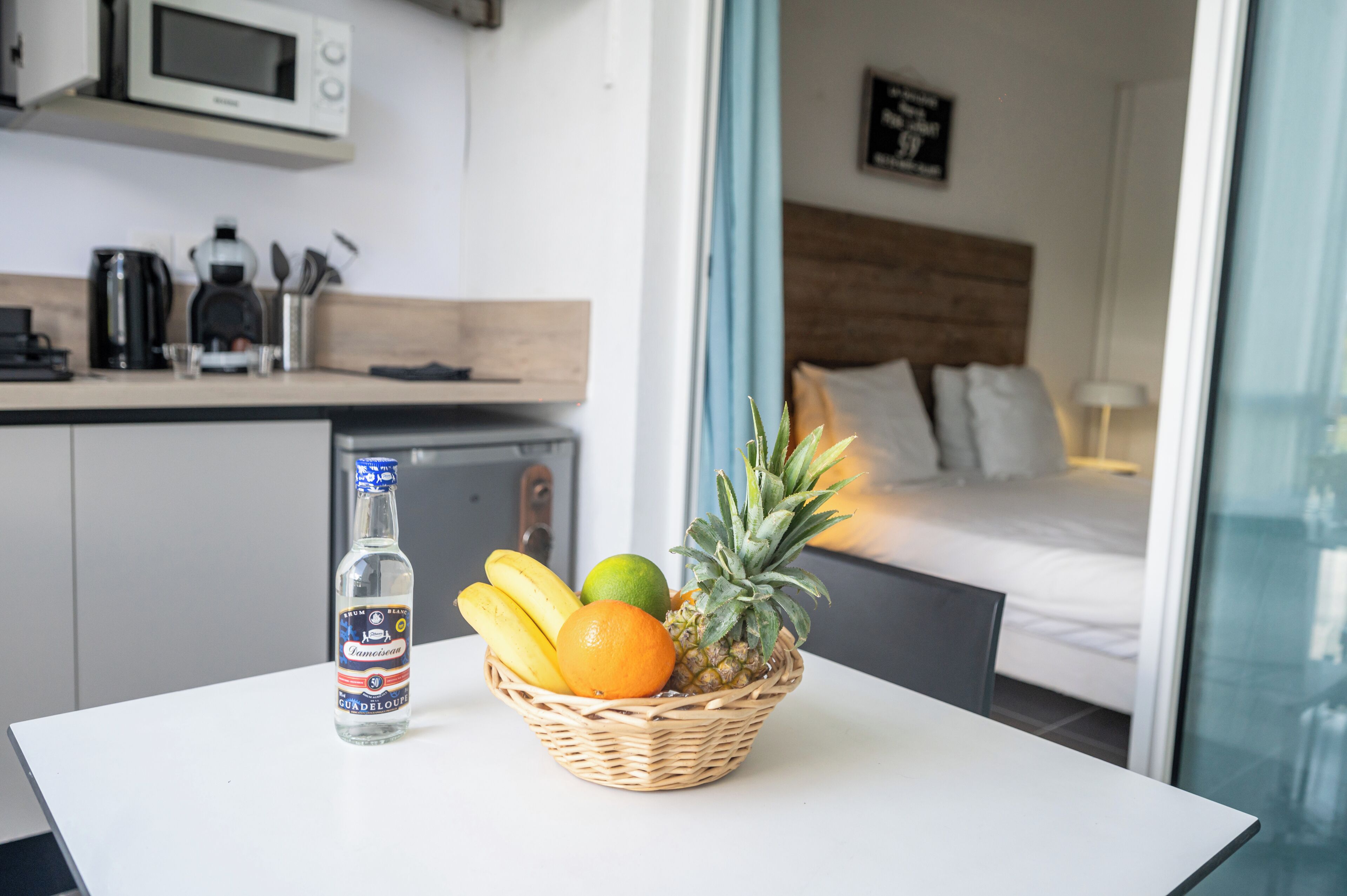 Une cuisine équipée d’un four à micro-ondes, d’une machine à café et d’ustensiles de cuisine. Un panier de fruits contenant des bananes, une orange et un ananas est posé sur une table. On peut également apercevoir une bouteille d’eau.
