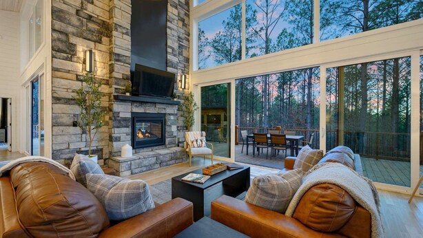 A modern living room with a stone fireplace, leather sofas, and large windows overlooking a forest.