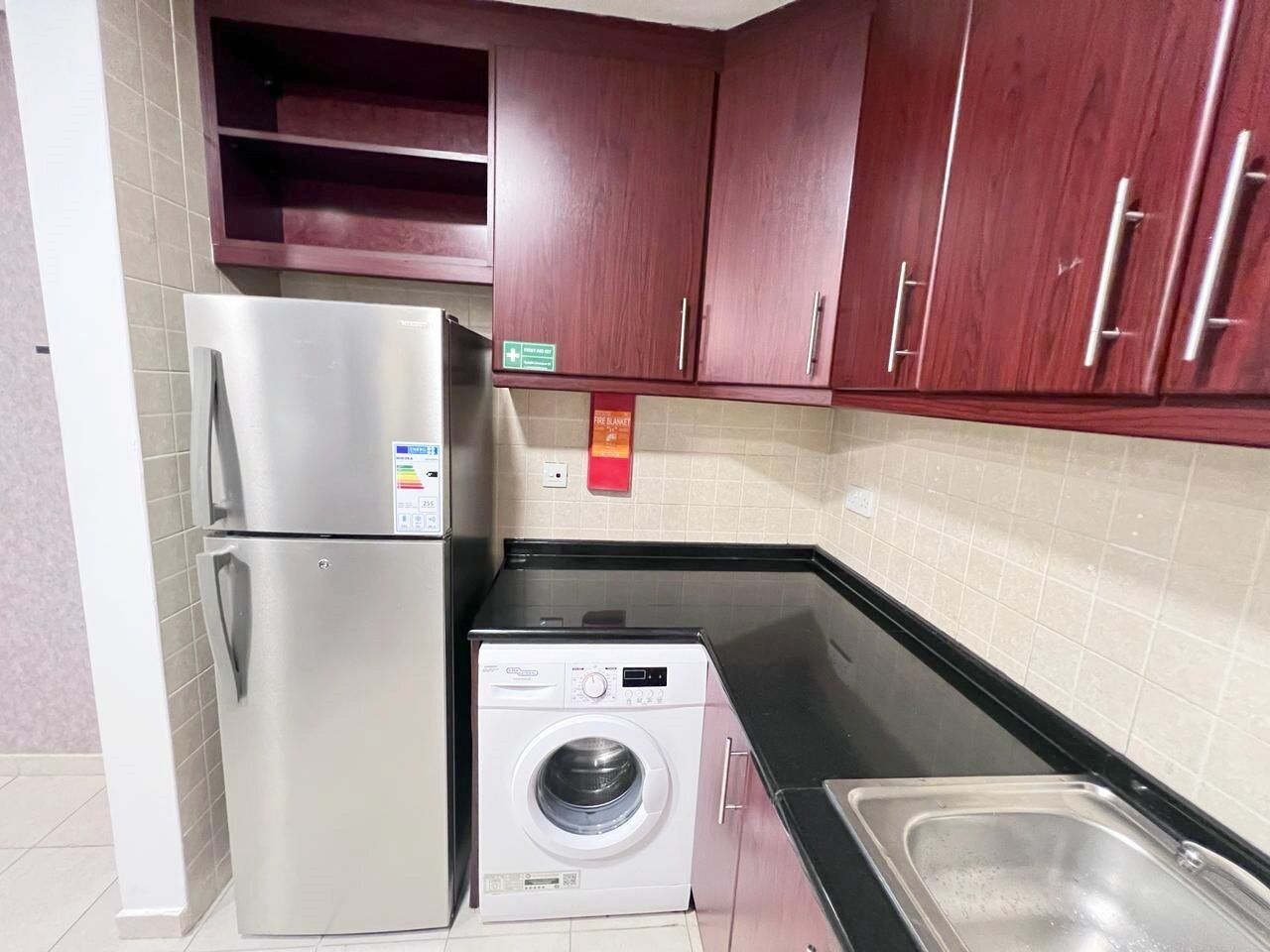Une cuisine compacte équipée d’un réfrigérateur en acier inoxydable, d’un lave-linge et de meubles de rangement en bois.