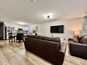 Una sala de estar moderna con un sofá de cuero marrón, un comedor con mesa de vidrio y una cocina al fondo.