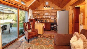 A cozy cabin interior with a wooden ceiling, a dining area with outdoor seating, a kitchen with wooden shelves, and a patterned rug. - Sunny Waterfront Cottage at FarAway Pond (Whitefield)