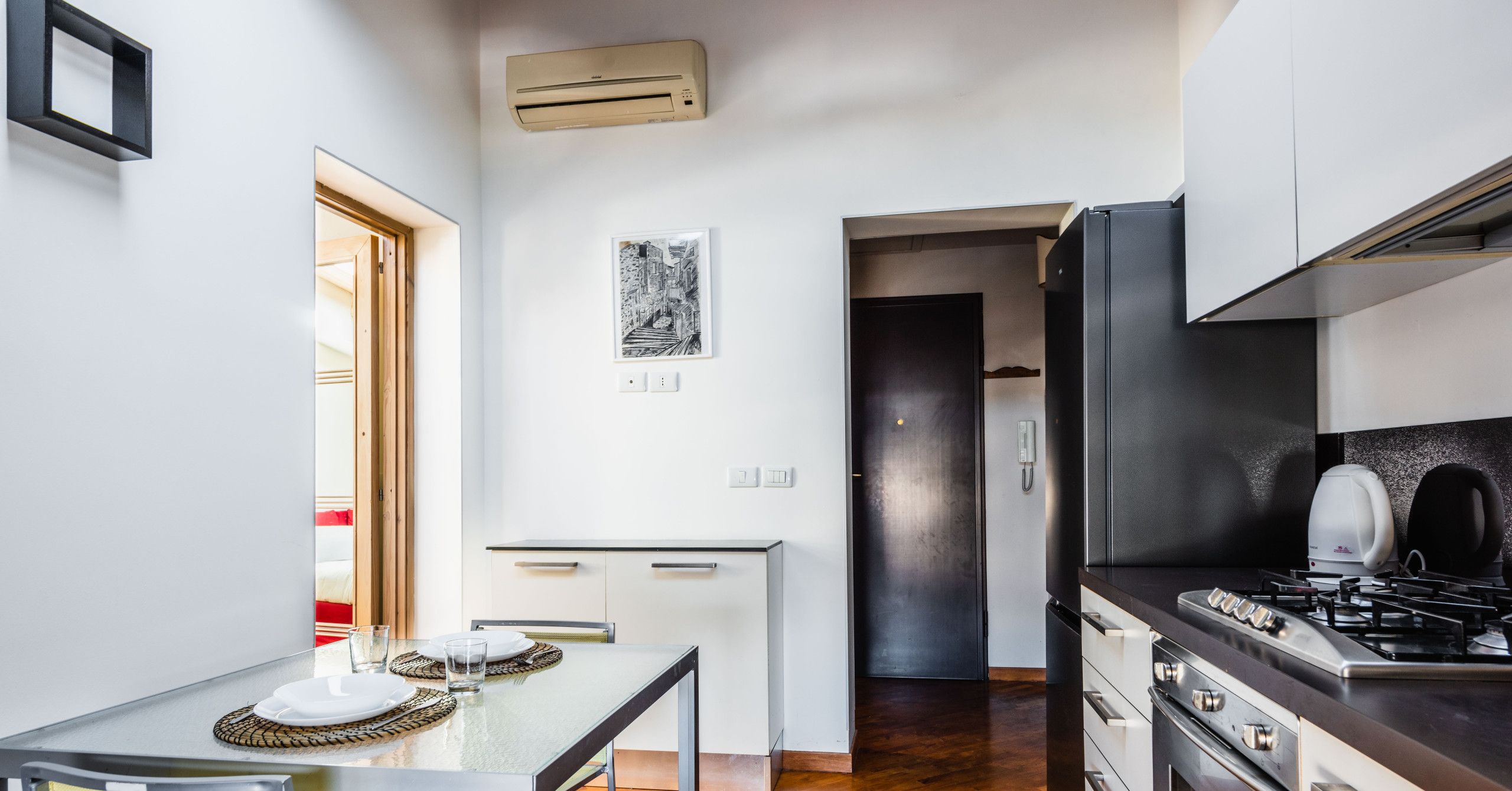 A modern kitchen with a dining area, featuring a glass table, stainless steel appliances, and a wooden floor.