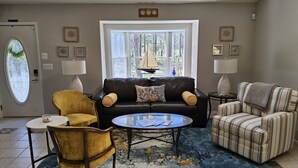 Un salon avec un canapé en cuir noir, un fauteuil jaune, un fauteuil à rayures, une table basse en verre et un tapis bleu.