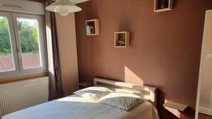 A bedroom with a bed, a nightstand, a window, and a wall-mounted shelf with books. - Ecully, Appartement au Calme de 50m² (Lyon)
