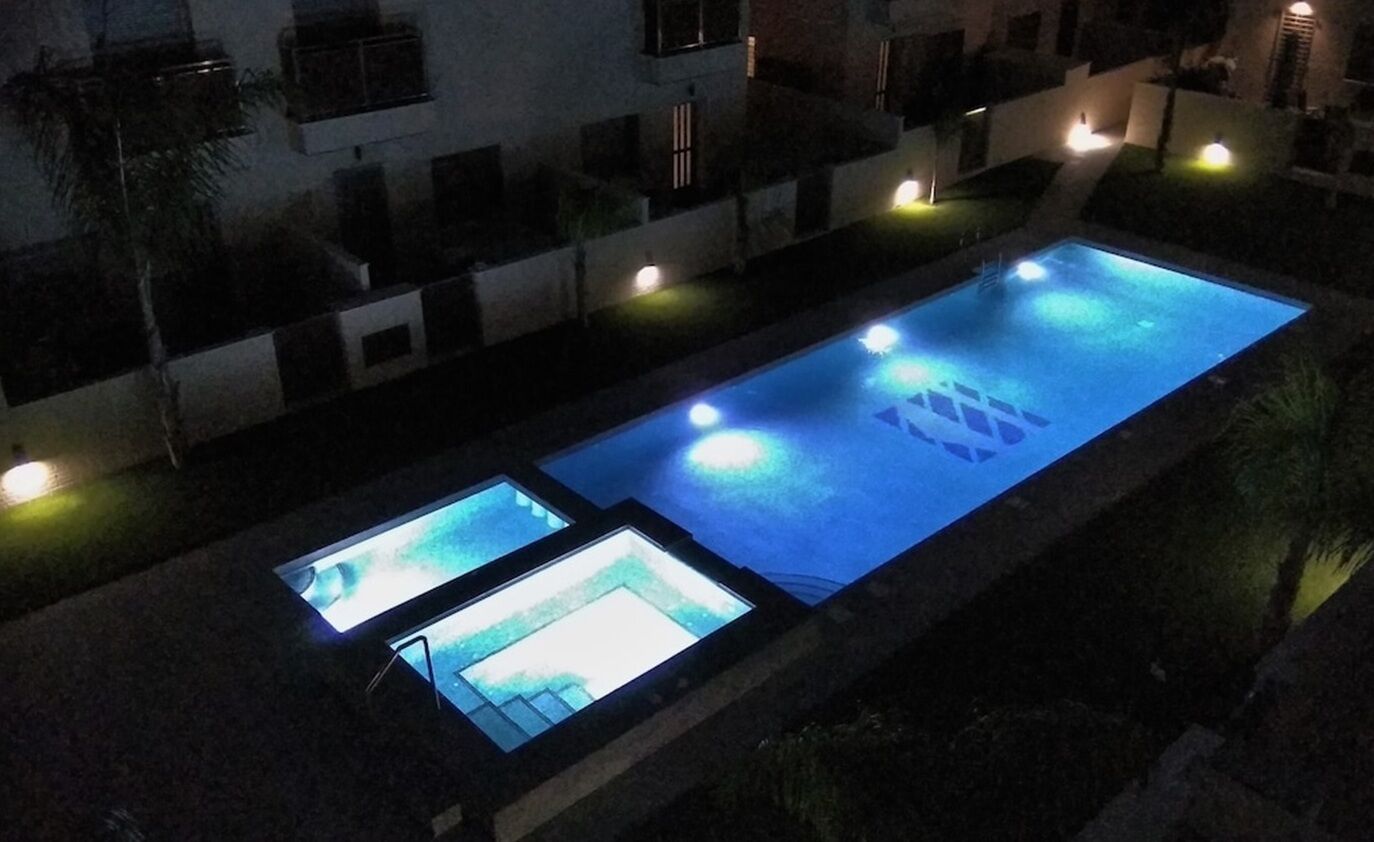 Piscina com fundo de vidro, rodeada por um edifício com várias varandas e iluminação.