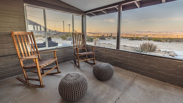 A room with two wooden rocking chairs and two knitted poufs on a concrete floor, overlooking a desert landscape through large windows.
