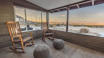 Une pièce avec deux fauteuils à bascule en bois et deux poufs en tricot, posés sur un sol en béton, d’où l’on peut admirer un paysage désertique à travers de grandes fenêtres.