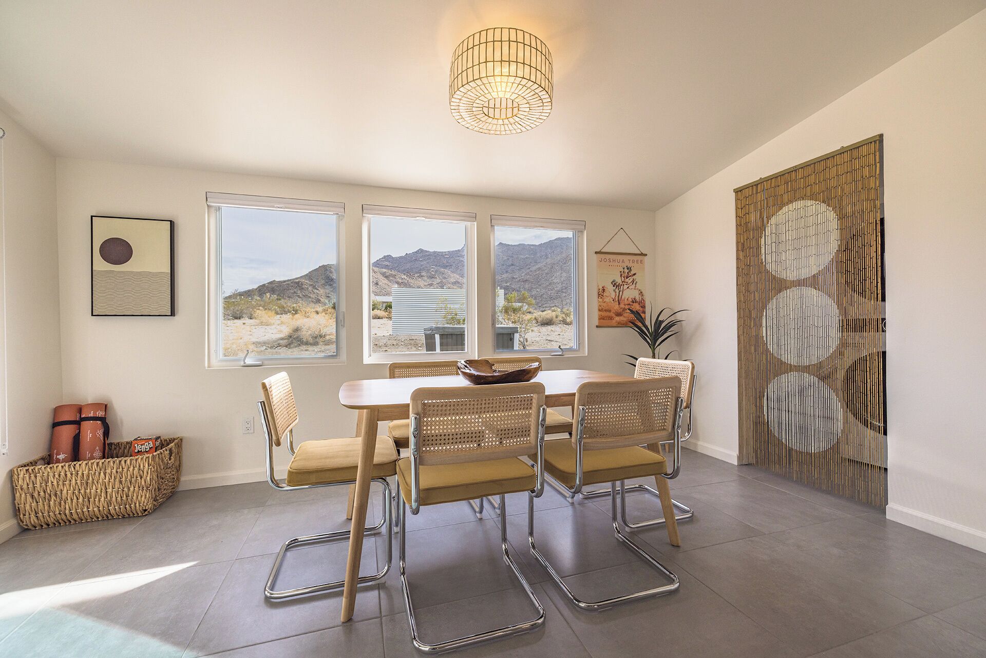 Un coin repas avec une table et quatre chaises, de grandes fenêtres donnant sur un paysage désertique, un luminaire suspendu et un panneau décoratif mural.