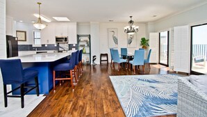 A modern kitchen with a central island, bar stools, and a dining area with blue chairs. - Summer Winds B-601, Oceanfront Condo in Indian Beach, NC! (Indian Beach)