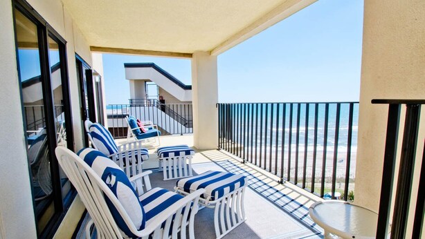 A balcony with white and blue striped chairs and a small table, overlooking a beach. - Summer Winds B-601, Oceanfront Condo in Indian Beach, NC! (Indian Beach)