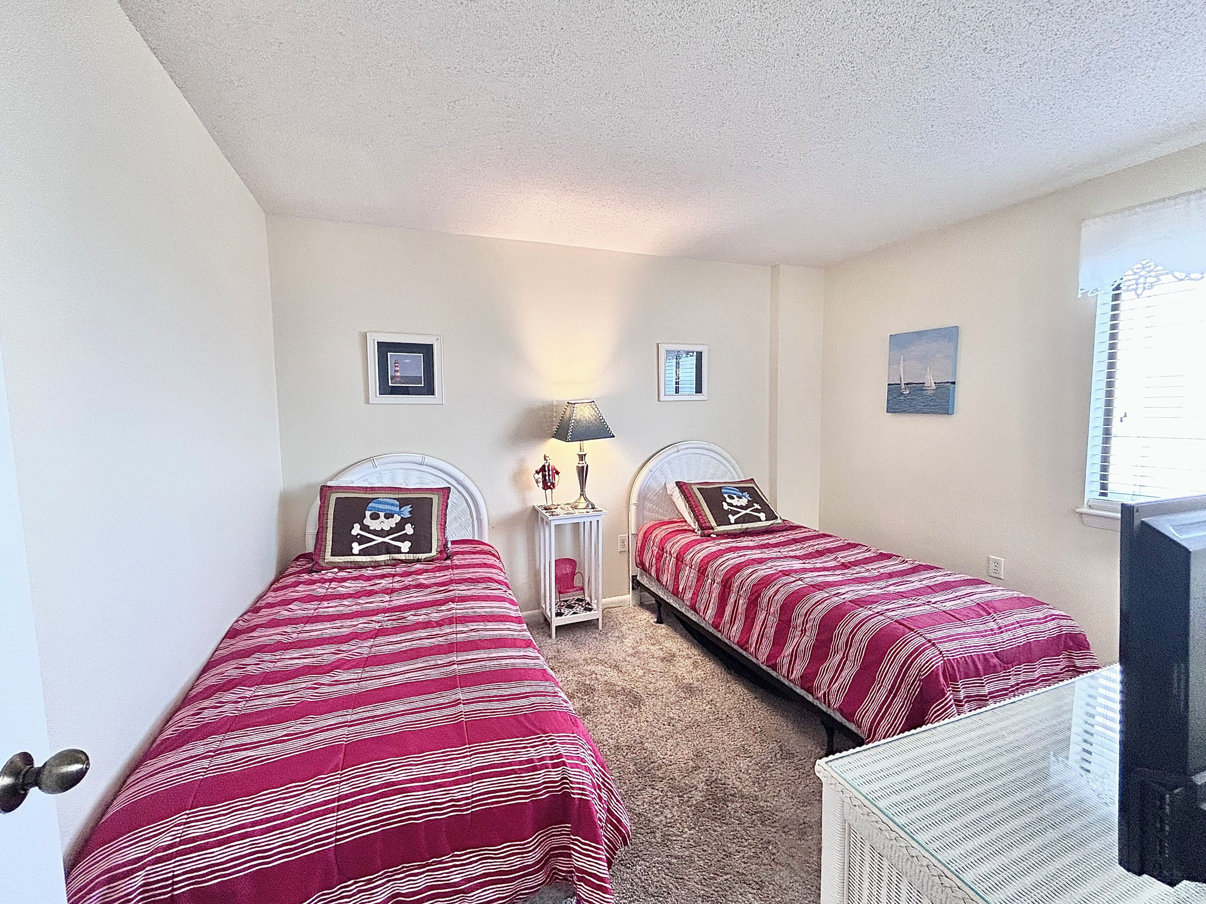 A bedroom with two single beds, a nightstand, a lamp, and a window with blinds.