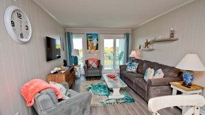 Un salon avec un canapé gris, un fauteuil blanc, une table basse et un téléviseur fixé au mur. Il y a deux fenêtres avec des rideaux, une horloge au mur et une petite étagère avec des objets décoratifs.