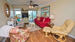 A living room with a red sofa, a beige chair, a television, and a painting of beach umbrellas. - SeaSpray 230, Oceanview Condo in Atlantic Beach, NC! (Atlantic Beach)
