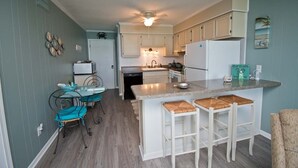 Une cuisine compacte avec des meubles blancs, un îlot central doté de tabourets, et un coin repas avec une table et des chaises en verre.