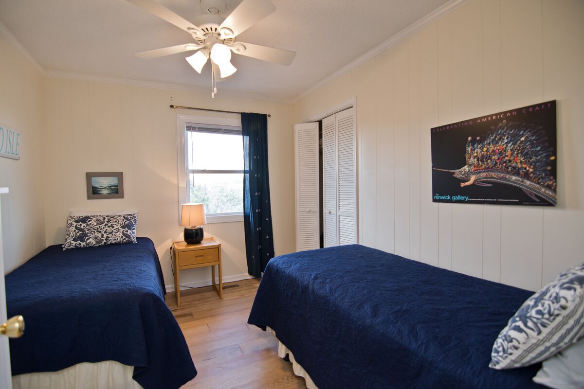 A bedroom with two beds, a ceiling fan, a window with curtains, a nightstand with a lamp, and a poster on the wall.