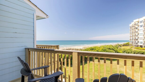 Een balkon met houten leuningen met uitzicht op een strand, een gebouw met een witte gevel en twee Adirondack-stoelen.