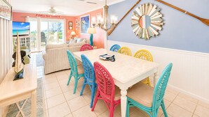 Uma sala de jantar com uma mesa branca e cadeiras coloridas, um ventilador de teto e um espelho decorativo na parede.