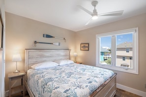 A bedroom with a bed, two bedside lamps, a ceiling fan, a window with a view, and a wall-mounted paddleboard. - Sound Of The Sea East 125, Oceanfront Condo in Emerald Isle, NC! (Emerald Isle)