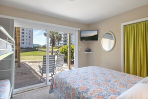 Una habitación de hotel con una cama, televisión, ventana con cortinas y puerta que da a un espacio exterior con sillas y vistas a edificios.