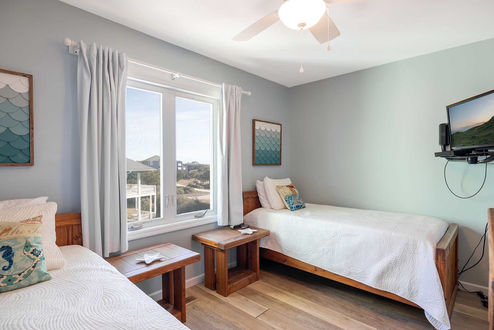 Une chambre avec deux lits, une fenêtre avec des rideaux, un ventilateur de plafond et un téléviseur fixé au mur.