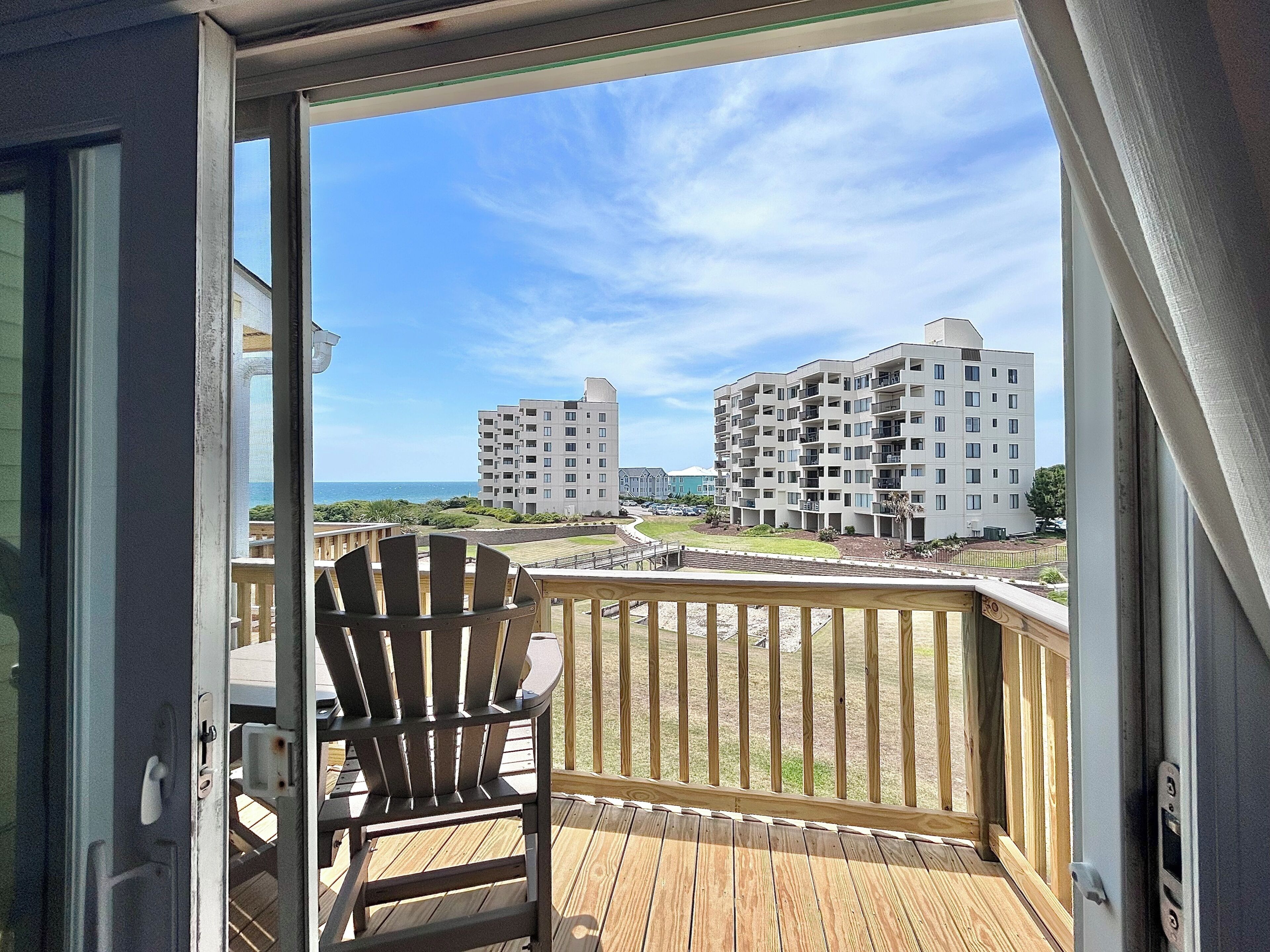 A balcony with a wooden deck, a Adirondack chair, and a view of multi-story buildings and a beach.