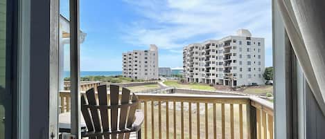 A balcony with a wooden deck, a Adirondack chair, and a view of multi-story buildings and a beach.