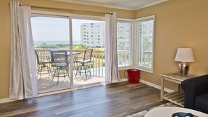 A room with a sliding glass door leading to a balcony, a small table with a lamp, and a sofa. - Sound Of The Sea North 205, Oceanview Condo in Emerald Isle, NC! (Emerald Isle)