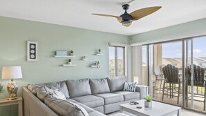 Una sala de estar moderna con un sofá seccional gris, una mesa de centro y un ventilador de techo. Hay estanterías con objetos decorativos y vistas al exterior a través de grandes puertas corredizas de vidrio.