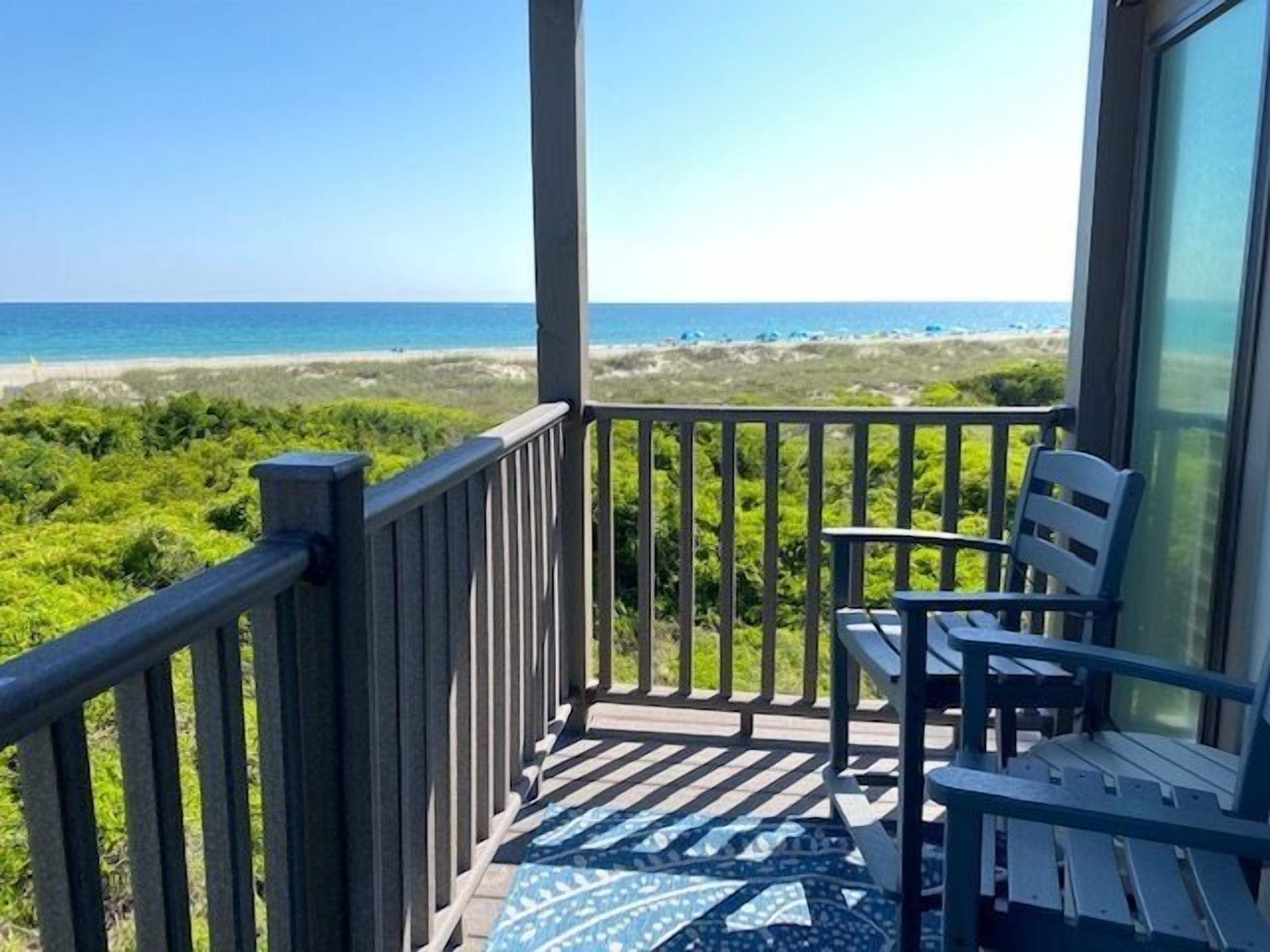 A balcony with a view of the beach, a chair, and a patterned rug.