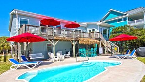 A pool area with lounge chairs and umbrellas, a wooden deck, and a building with a balcony. - Stay Salty, Almost Oceanfront House in Emerald Isle, NC! (Emerald Isle)