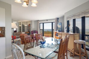 A spacious living area with a glass-top dining table, comfortable seating, and a large window offering a view of the beach. - Summer Winds C-120, Oceanfront Condo in Indian Beach, NC! (Indian Beach)
