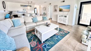 A living room with a sofa, coffee table, and a TV. There is a kitchen in the background. - Summer Winds C-128, Oceanfront Condo in Indian Beach, NC! (Indian Beach)