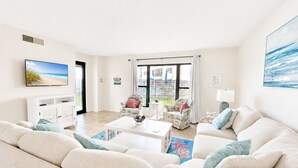 A spacious living room with a large sofa, a coffee table, and a television. There is a balcony with a view of the beach. - Summer Winds C-128, Oceanfront Condo in Indian Beach, NC! (Indian Beach)