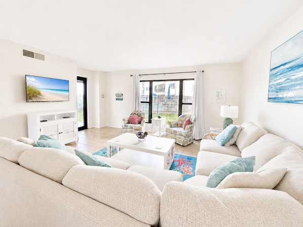 A spacious living room with a large sofa, a coffee table, and a television. There is a balcony with a view of the beach.