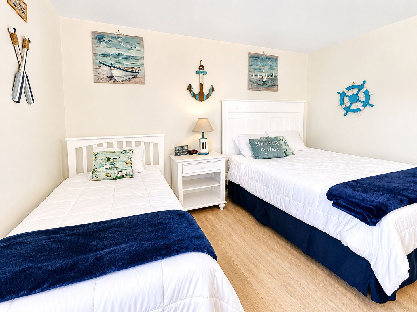 A bedroom with two beds, a nightstand, a lamp, and nautical-themed wall art.