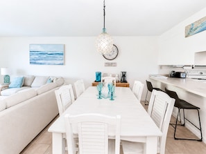 Une cuisine moderne avec des meubles blancs, un coin repas avec une table et des chaises blanches, et un salon avec un canapé et un tableau représentant l’océan.