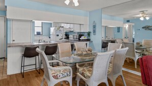 Une cuisine moderne avec des meubles blancs, un îlot central et un coin repas comprenant des chaises en osier et une table en verre.