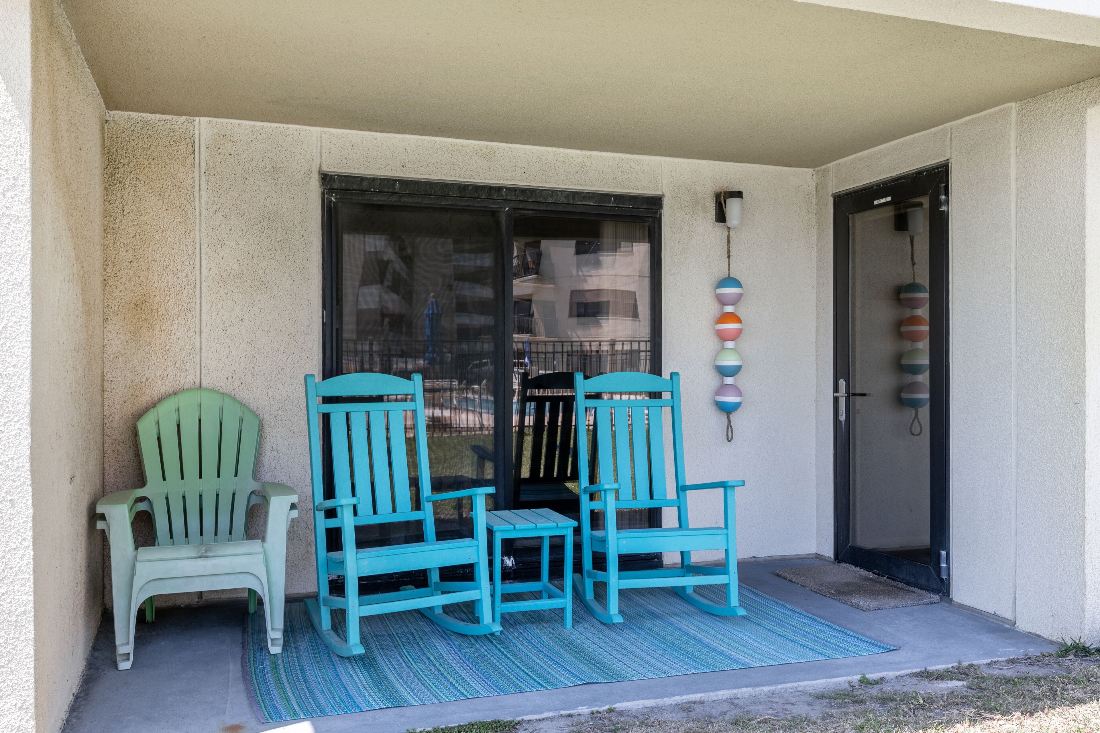 Un balcon avec deux chaises turquoise et une petite table, une porte noire et un élément décoratif fixé au mur.
