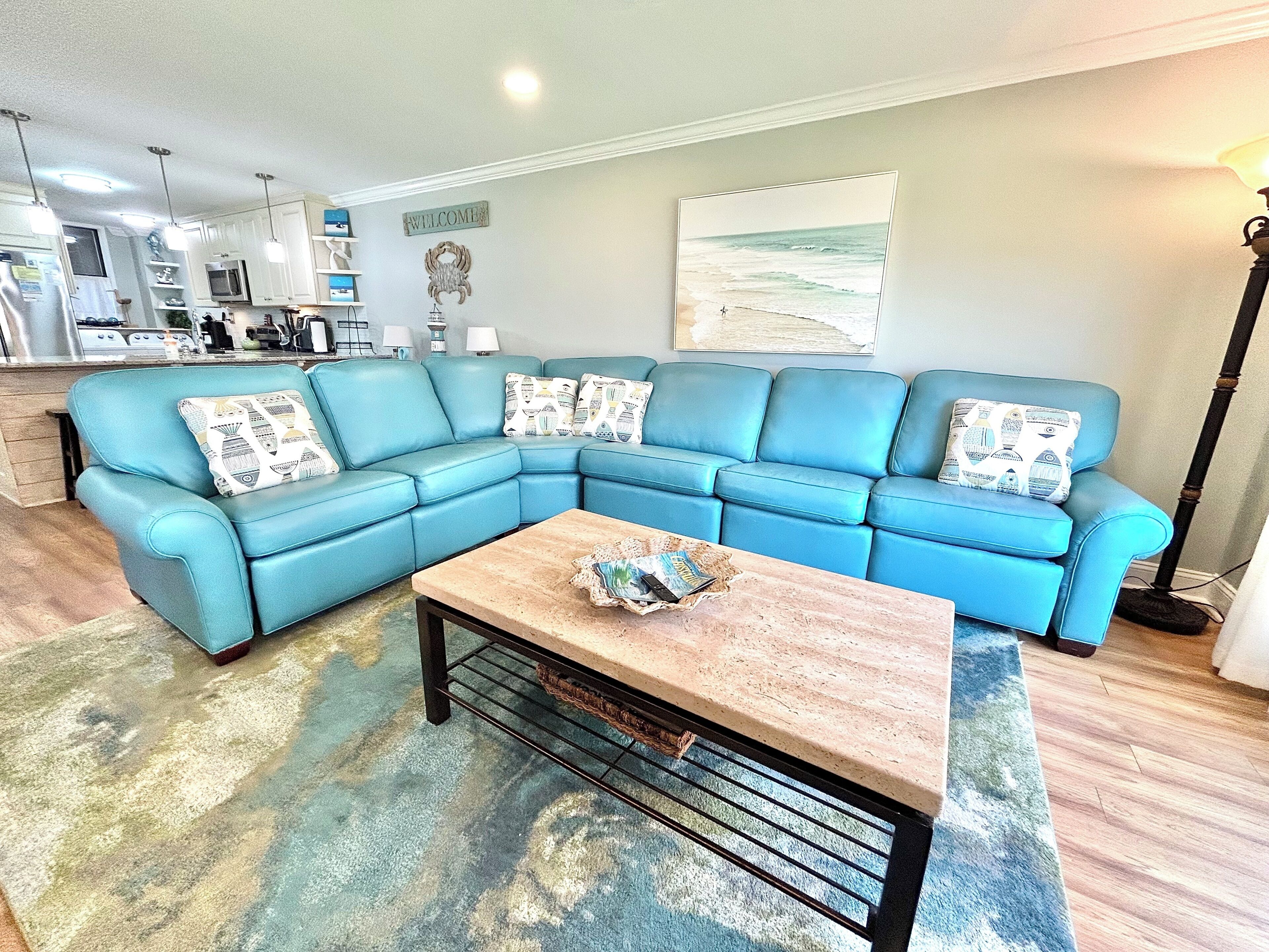 A living room with a blue sectional sofa, a coffee table, and a kitchen in the background.