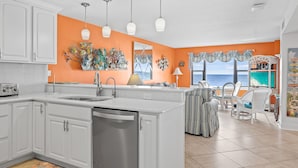 A kitchen with white cabinets, a stainless steel dishwasher, and a large window overlooking the ocean. - Summer Winds 221, Oceanfront Condo in Indian Beach, NC! (Indian Beach)