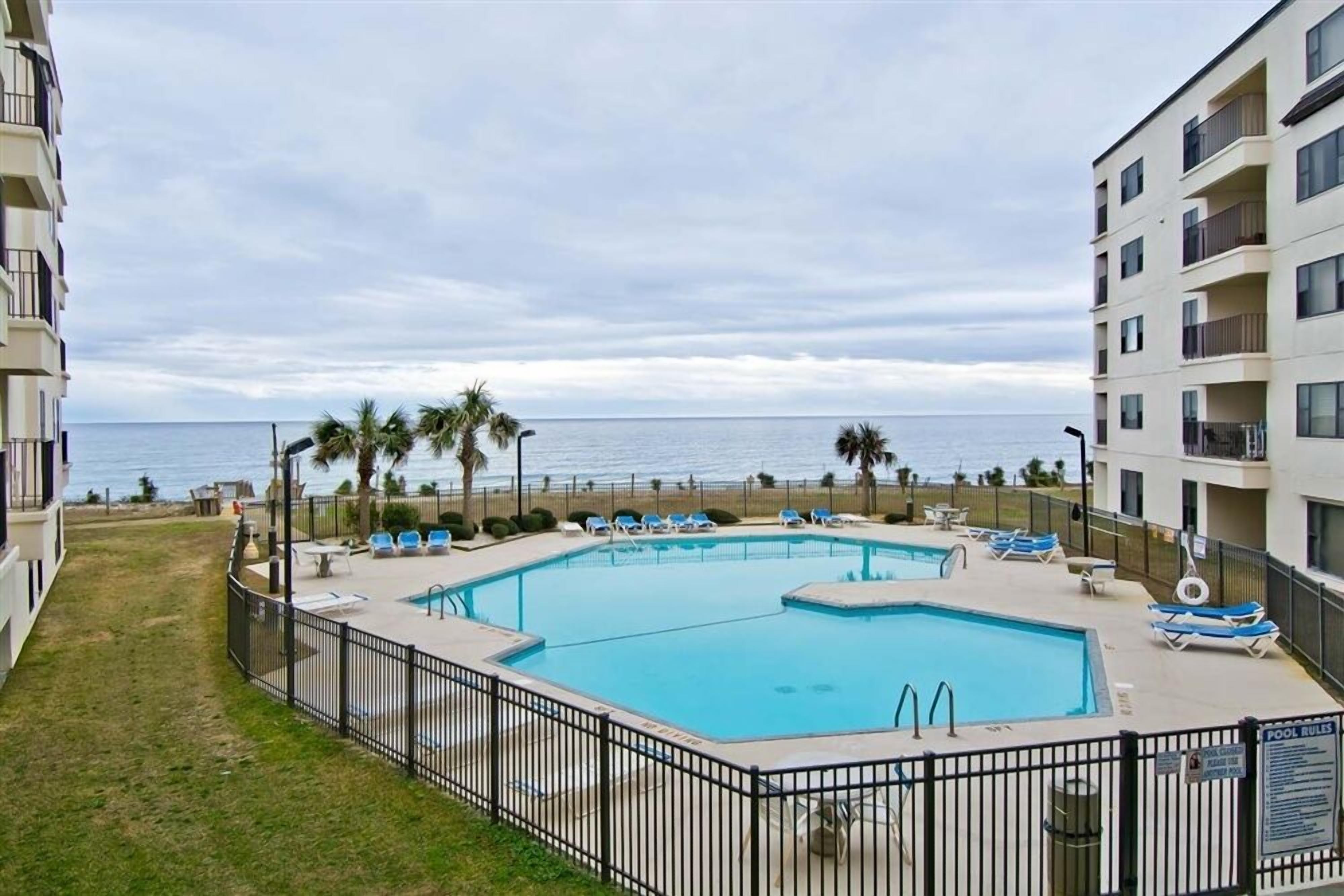 A rectangular swimming pool with a black fence, surrounded by lounge chairs and palm trees, overlooking the ocean.