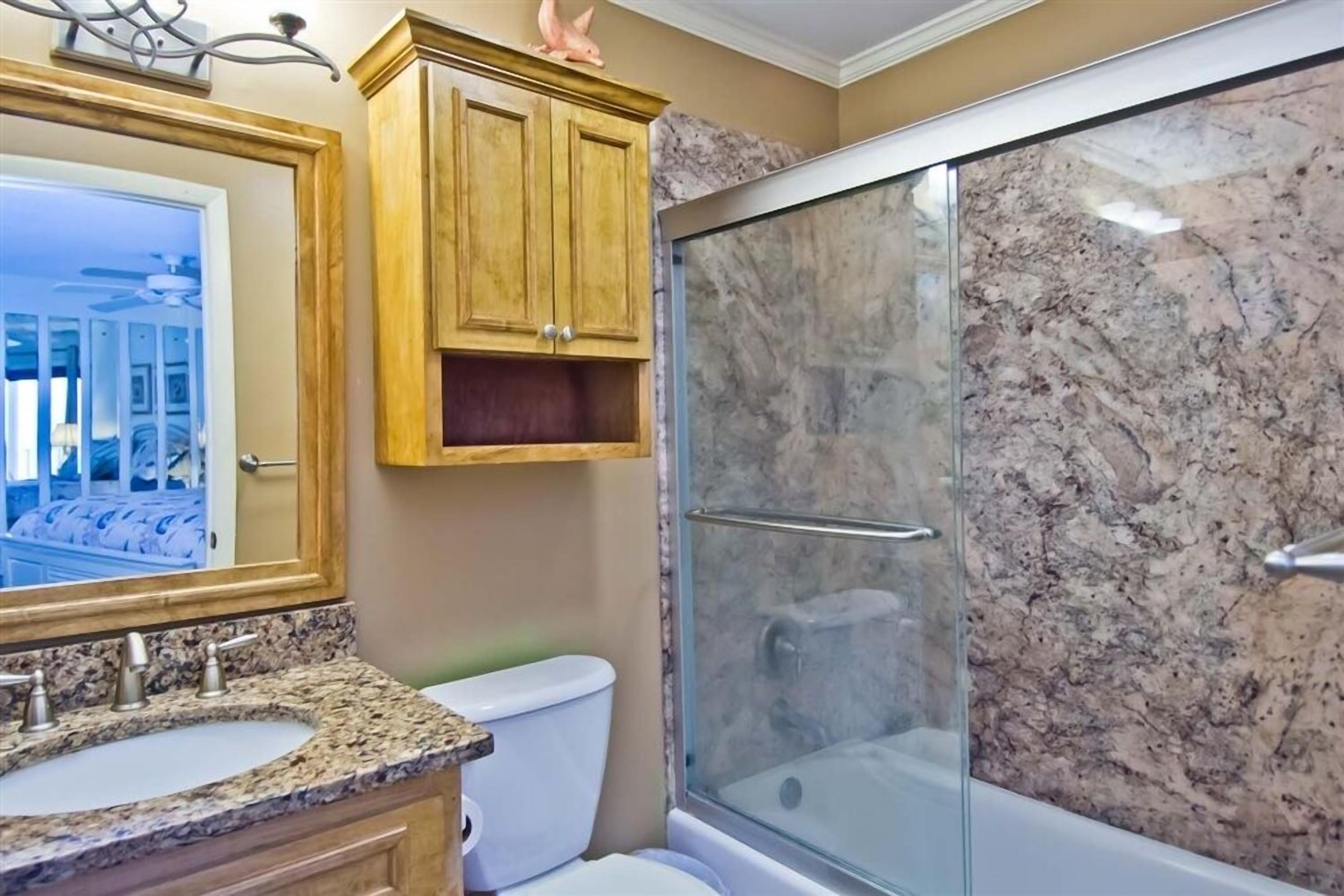 A bathroom with a granite countertop sink, wooden cabinets, a toilet, and a glass-enclosed shower.