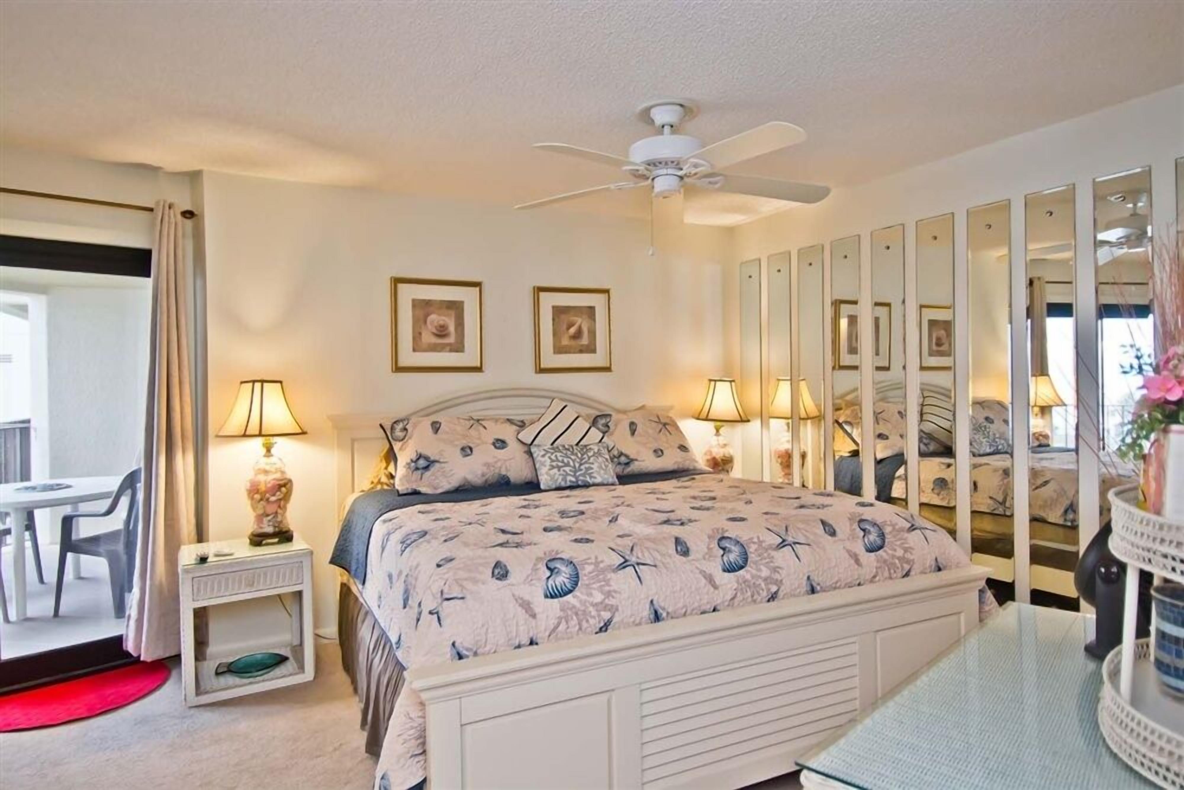 Une chambre à coucher avec un lit, des lampes de chevet, un ventilateur de plafond, un miroir et une petite table sur laquelle est posé un vase.