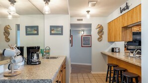 A kitchen with granite countertops, wooden cabinets, a coffee maker, and a toaster. There are framed pictures on the wall and a sign that reads "FISHING TRIPS". - Summer Winds B-237, Oceanfront Condo in Indian Beach, NC! (Indian Beach)