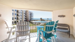 A balcony with white and blue Adirondack chairs, a wicker sofa, and a view of the ocean and a building with scaffolding. - Summer Winds B-237, Oceanfront Condo in Indian Beach, NC! (Indian Beach)