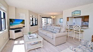A living room with a sofa, coffee table, TV, and a dining area with chairs and a table. - Summer Winds A-301, Oceanfront Condo in Indian Beach, NC! (Indian Beach)