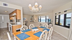 A modern kitchen with wooden cabinets, a dining area with a wooden table and white chairs, and a view of the sea through large windows. - Summer Winds A-301, Oceanfront Condo in Indian Beach, NC! (Indian Beach)