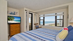 A bedroom with a bed, a television, a wooden dresser, a chair, and a window with a beach view. - Summer Winds A-301, Oceanfront Condo in Indian Beach, NC! (Indian Beach)